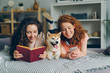 © silverkblack - Girls friends are reading book and using smartphone lying on bed with cute shiba inu dog in cozy apartment. Modern lifestyle, friendship and animals concept.