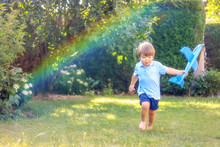 Cute Little Boy Playing With Toy Plane Model Running Barefoot On Grass In Garden With Rainbow Lens Dispersion Defect . Happy Carefree Childhood, Summer Lifestyle.