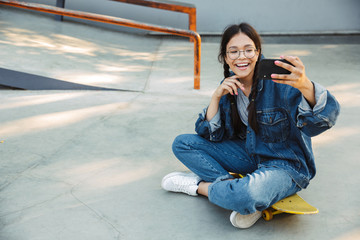 Poster - Image of cute girl taking selfie photo on smartphone while sitting on skateboard in skate park