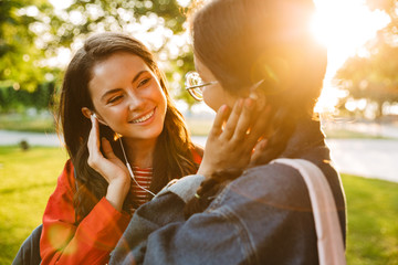 Poster - Image of two cute girls students using earphones and looking at each other while walking in green park