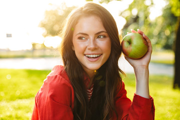 Wall Mural - Image of lovely girl student jacket smiling and holding apple while sitting in green park