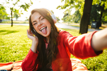 Sticker - Image of funny girl with tongue sticking out using headphones and taking selfie photo while sitting in green park