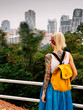 © Artem Markin - The blonde stands with his back to the camera, looking at the city from a high point. Wearing white jersey, blue skirt, orange backpack. Hands with tattoos. Asia travel