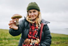 Young Beautiful Woman In Camping Outdoor Outfit For Cold Harsh Weather Holds Out Wild Fresh Picked Mushroom To Camera. Concept Healthy Eating And Local Organic Produce Farm