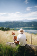 © BublikHaus - Young male hiker or adventurer tourist takes rest at scenic overlook at field on top of mountain together with pet dog basenji brown puppy. Healthy lifestyle and outdoor activities