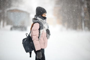 Wall Mural - Young cute white caucasian girl in pink down jacket, cap, warm scarf and gloves with a backpack stands on the street in winter.