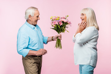 Wall Mural - cheerful senior man giving bouquet with flowers to excited wife on pink