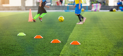 selective focus to marker cones are soccer training equipment on green artificial turf with blurry kid players training background. material for training class of football academy.