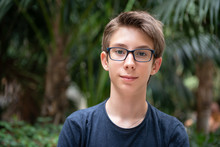 Young Boy Posing In Summer Park With Palm Trees. Cute Spectacled Smiling Happy Teen Boy 13 Years Old, Looking At Camera. Kid's Outdoor Portrait.