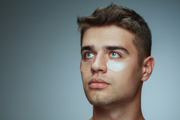 Wall Mural - Close-up profile portrait of young man isolated on grey studio background. Male face with collagen patches under eyes. Concept of men's health and beauty, cosmetology, body and skin care. Anti-aging.