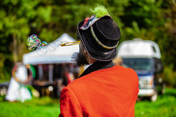 Fusion of cultural & modern music event. A shaman is viewed from the back, in traditional costume, blowing soap bubbles in a campsite at a music festival, blurry RV and tent is seen in background