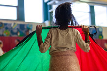 Kindergarten Children Educational Game. A Closeup And Rear View Of A Young Black Girl Holding A Rainbow Parachute During A Physical Education Lesson In A School Gymnasium