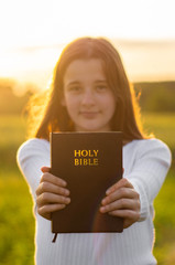 Wall Mural - Christian teenage girl holds bible in her hands. Reading the Holy Bible in a field during beautiful sunset. Concept for faith, spirituality and religion. Peace, hope