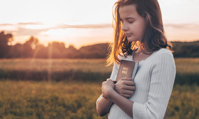 christian teenage girl holds bible in her hands. reading the holy bible in a field during beautiful 