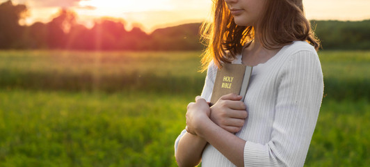 Wall Mural - Christian teenage girl holds bible in her hands. Reading the Holy Bible in a field during beautiful sunset. Concept for faith, spirituality and religion. Peace, hope