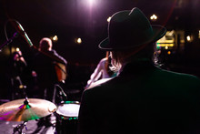 Musicians Entertain People In Night Club. A Rear View Of A Silhouetted Man Wearing A Top Hat As He Performs A Music Gig On Stage With Fellow Band Members In The Background And Copy Space On The Left.