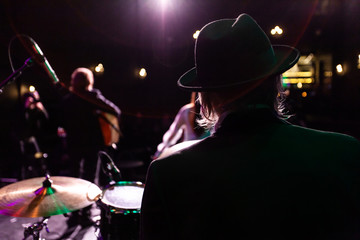 Musicians entertain people in night club. A rear view of a silhouetted man wearing a top hat as he performs a music gig on stage with fellow band members in the background and copy space on the left.