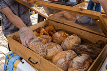Organic Produce Sold At Farmer's Market. Hands Of A Baker Are Seen Reaching Into A Display Cabinet Filled With Loaves Of Freshly Baked Bread On A Stall During A Fair For Local Food Producers.