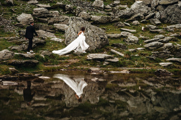 Wall Mural - Side view portrait of a amazing bride running into mountains near a lake while her groom is walking