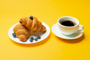 French croissants, cup of coffee and blueberries on yellow background close up	