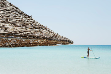 Beautiful beach resort with straw umbrellas on a blue sky and white clouds. On the background a man is surfing standing up with a paddle. Beach life and lifestyle concept.