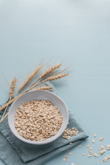 Wall Mural - Bowl of dry oat flakes with ears of wheat on light blue background. Cooking oats porridge concept