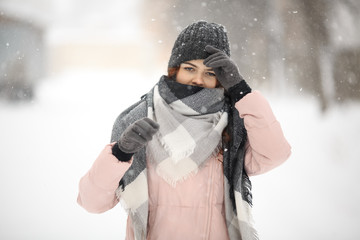 Wall Mural - Young cute white caucasian girl in pink down jacket, cap, warm scarf and gloves stands on the street in winter.