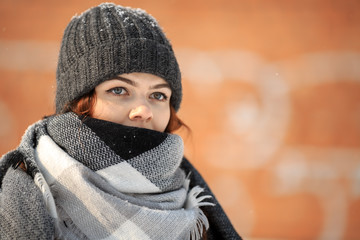 Wall Mural - A young cute white girl in a hat and warm scarf is standing in the street opposite red background in winter. Close up.