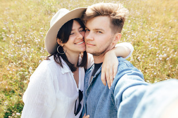 Happy hipster couple making selfie in sunny wildflower meadow, traveling in mountains. Stylish young family smiling and enjoying hiking on top of mountain, taking photo. Summer vacation