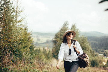 Wall Mural - Happy hipster girl with backpack traveling on top of sunny mountain, walking on hills with woods. Stylish woman smiling and  enjoying hiking in mountains. Wanderlust and travel.