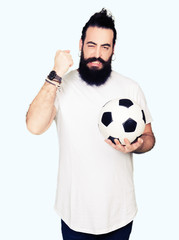 Wall Mural - Young man with long hair and bear holding soccer football ball annoyed and frustrated shouting with anger, crazy and yelling with raised hand, anger concept