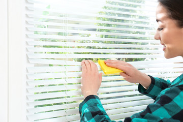 Canvas Print - Beautiful woman cleaning window blinds with rag indoors