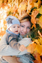 Wall Mural - Happy mother with her lovely baby lying in autumn leaves outdoor. Cheerful woman hugs her son.