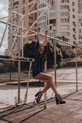 Fashion young beauty woman with blonde hair in classic sundress and heels posing on gate on sports ground.