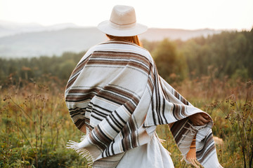 Wall Mural - Stylish boho girl walking in sunny light at atmospheric sunset in meadow. Happy hipster woman in poncho and hat enjoying traveling in evening mountains. Space for text