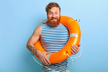 Glad man rescuer poses with orange lifebuoy, wears striped sailor vest, has red hair and beard, rests during summer holiday, has tattoo on arm, isolated on blue background, ready for swimming