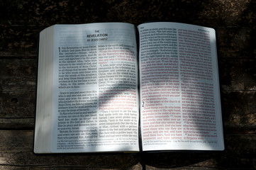 Open Bible in Revelation of Jesus Christ in chapters 1 and 2 with sunshine in the texts. Background with wooden table.