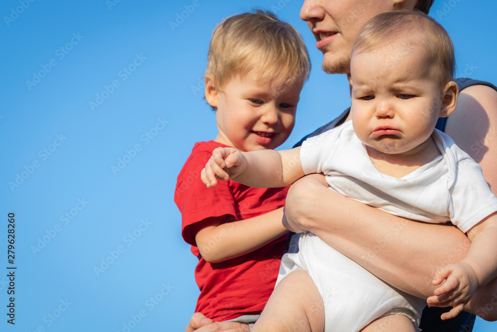 Young Father holds two children against the blue sky. Toddler is happy and laughing, and the baby is crying and sad. The concept of family, jealousy, relationships, sisters and brothers. Copy space