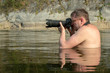 A professional photographer with glasses sits on a mountain river in front of his chest and holds a digital camera in his hands. A photo hunter or a reporter journalist of old agework in the water
