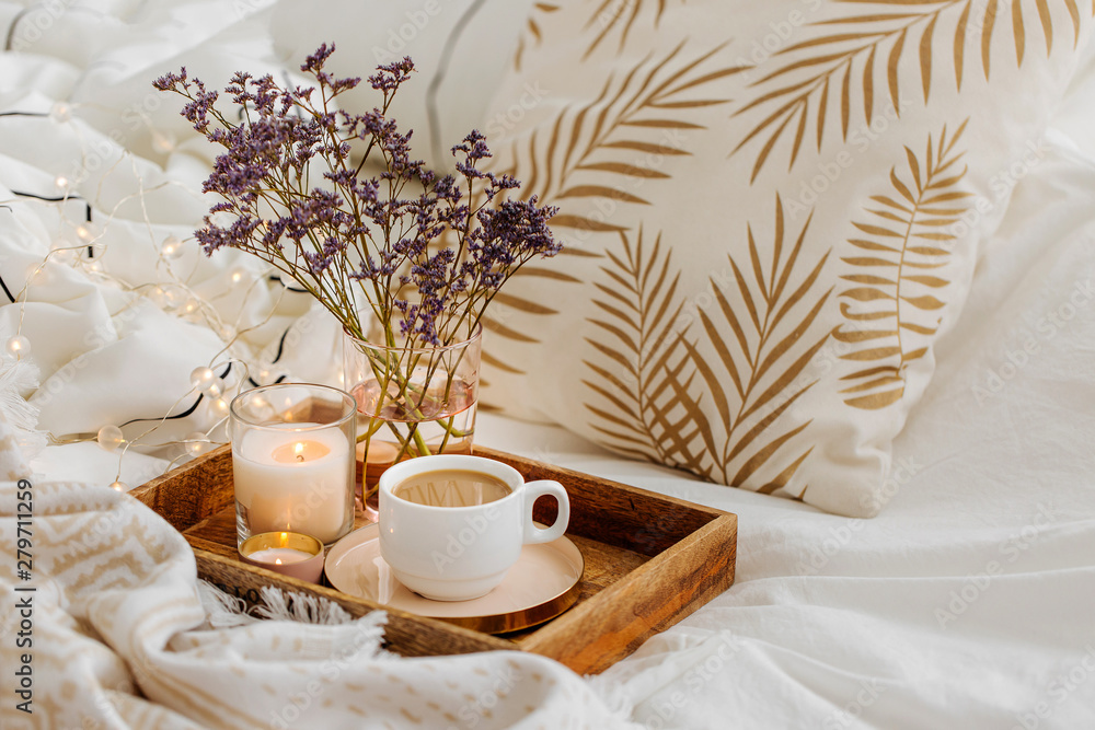 Wooden tray of coffee and candles with flowers on bed. White bedding sheets with striped blanket and pillow. Breakfast in bed. Hygge concept. - obrazy, fototapety, plakaty 