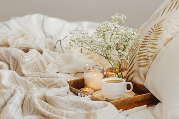 Wooden tray of coffee and candles with flowers on bed. White bedding sheets with striped blanket and pillow. Breakfast in bed. Hygge concept.