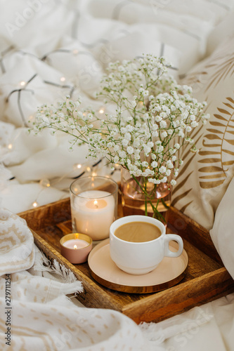 Wooden tray of coffee and candles with flowers on bed. White bedding sheets with striped blanket and pillow. Breakfast in bed. Hygge concept.