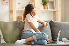 Content modern young woman in casual outfit sitting on sofa and using laptop while making order online and calling website operator