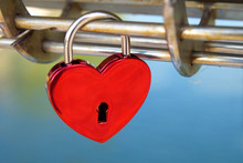 New Heart-shaped Red Love Lock Hanging On A Metal Bridge. Lock On Partially Rusty Nickel-plated Rod. Gloss Padlock With Keyhole On Blurred Blue Water Background. Saint Valentines Day Card Background.