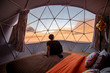 © zephyr_p - Asian tourist man sitting on the bed in dome tent looking outside at Wadi Rum desert, famous natural attraction in Jordan. Travel Middle east concept
