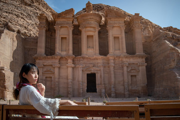 Asian woman tourist in white dress sitting and looking at Ad Deir or El Deir, the monument carved out of rock in the ancient city of Petra, Jordan. Travel UNESCO World Heritage Site in Middle East