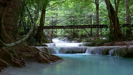 Wall Mural - Wooden bridge over the Erawan cascade waterfall. Kanchanaburi National Park, Thailand. Amazing tourist attraction. Wonderful destination. Fairytale atmosphere. Lush tropical vegetation, ancient trees