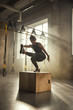 © Friends Stock - Becoming the best. Side view of athletic woman in sportswear doing squat and training legs while standing on wooden box at gym.