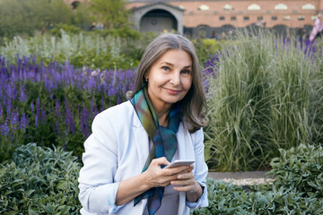 Wall Mural - Electronic gadgets, age, networking and communication concept. Outdoor image of blue eyed wrinkled woman in fashionable elegant clothes messaging online using mobile phone, smiling at camera