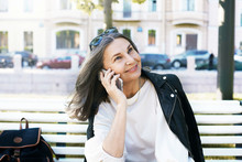 Adorable Charming Fifty Year Old Caucasian Female With Stylish Shades On Her Head Relaxing In City Surroundings, Sitting On Bench With Backpack, Talking On Mobile Phone. Mature Woman Calling Friend
